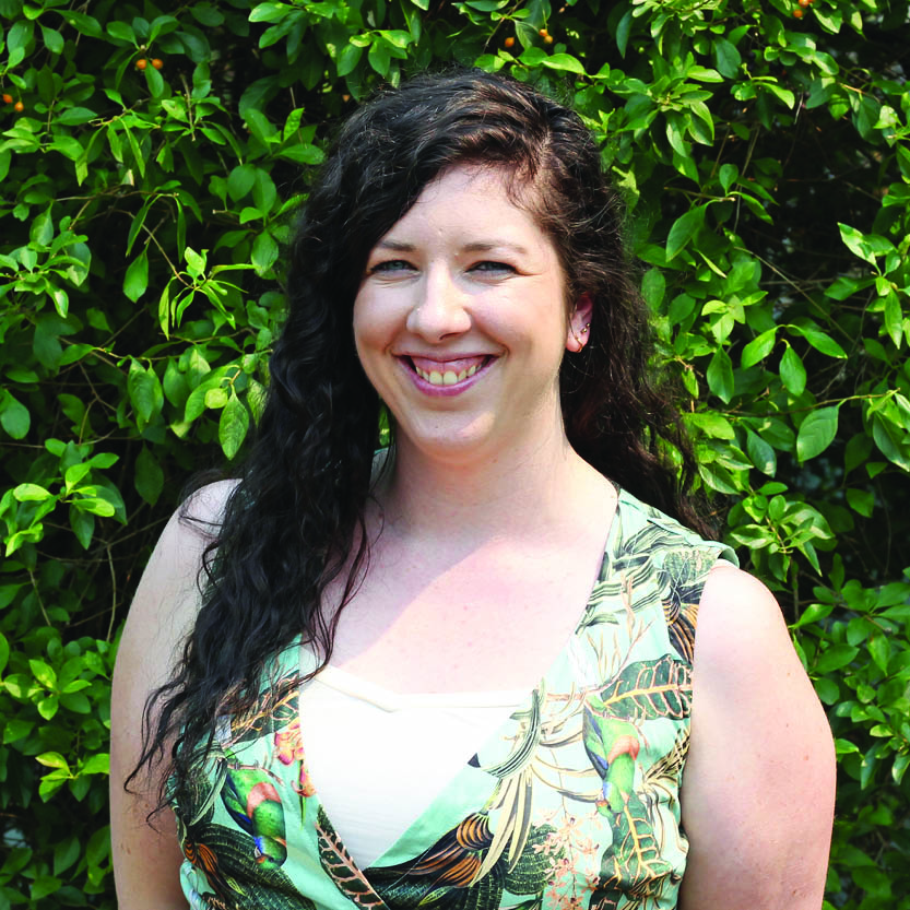 Image of lady with brown curly hair, smiling and standing in front of green trees